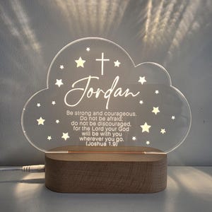 May include: A clear acrylic cloud-shaped night light with the name "Jordan" and a cross design. The light includes the text "Be strong and courageous..." and star accents. It sits on a wooden base.