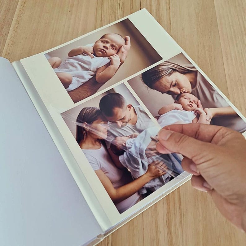 May include: A photo album with self-adhesive pages featuring three printed photographs of a baby and family members. The album has a white border and a clear plastic film overlay that is being lifted to reveal the glossy color photographs inside.