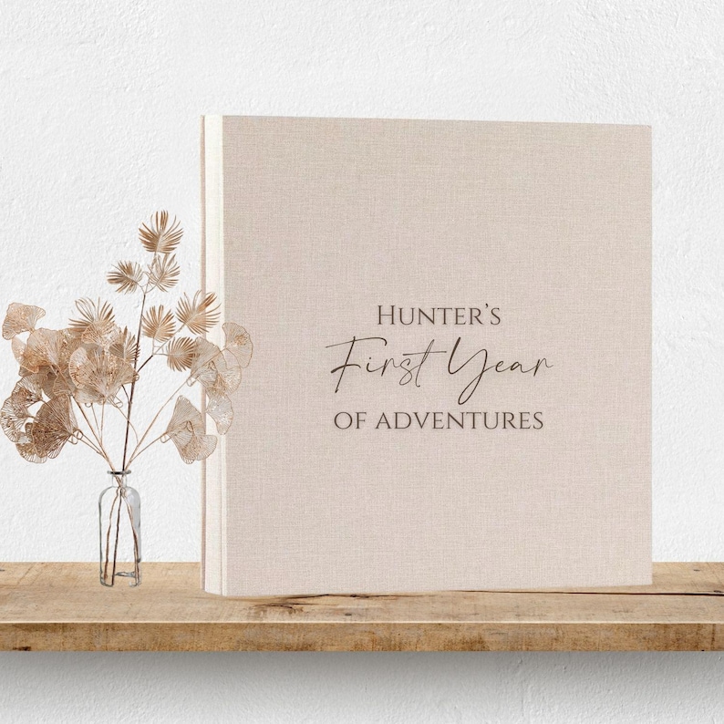 May include: A square photo album with a light beige linen cover. The front cover features the dark brown printed text "HUNTER'S First Year OF ADVENTURES" in a combination of serif and script fonts. The album is positioned next to a clear glass vase.