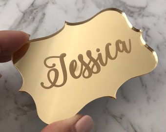 May include: Gold mirror place card with the name "Jessica" engraved in a cursive font.