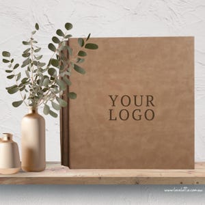 May include: A brown leather-bound book with the text "YOUR LOGO" embossed on the cover. The book is displayed on a wooden shelf with a gold vase and greenery. The website address www.lovelottie.com.au is visible.
