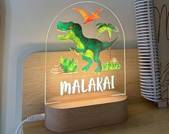 Puede incluir: Una luz nocturna de madera con un inserto de acrílico transparente que presenta un dinosaurio verde, un pterodáctilo naranja volador y palmeras verdes. El nombre "MALAKAI" está escrito en letras blancas debajo de la imagen.