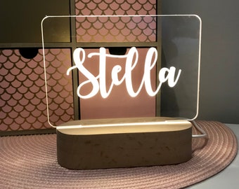 May include: A white acrylic sign with the name "Stella" illuminated with white LED lights. The sign is sitting on a wooden base and is on a pink and white rug.