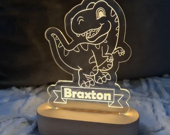 May include: A white acrylic night light shaped like a cartoon dinosaur with a banner that reads "Braxton". The night light is lit up with a warm yellow glow and sits on a wooden base.