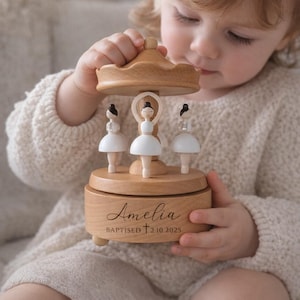 May include: A wooden carousel music box featuring three ballerina figurines in white tutus. The box is engraved with the name "Amelia" and the text "BAPTIZED 2.10.2025". The carousel has a light brown finish.