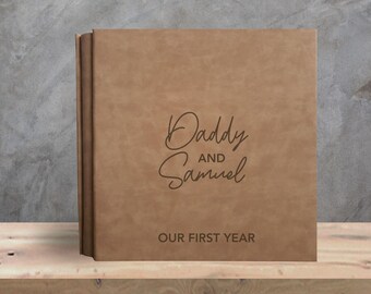 May include: A brown, leather-bound photo album with the words "Daddy and Samuel" and "Our First Year" embossed on the cover. The album is displayed on a wooden shelf against a gray background. The text "Text Design 1" is at the bottom.