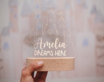 Puede incluir: Una luz nocturna de acrílico blanco con forma de castillo con el nombre Amelia y el texto "Dreams Here" inscrito en ella. La luz nocturna está sentada sobre una base de madera.
