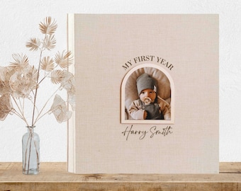 May include: A beige linen-covered photo album with a cutout arch shape. The arch contains a photo of a baby wearing a gray hat and blue overalls. The text "My First Year" is above the photo and "Harry Smith" is below the photo.