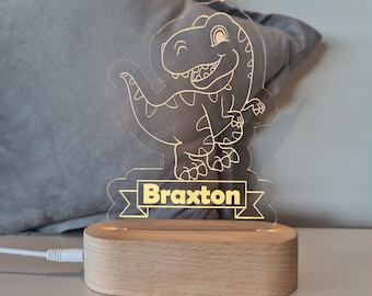 Könnte beinhalten: Ein Holzsockel mit einem klaren Acryl-Dinosaurier-Nachtlicht. Der Dinosaurier ist in Gelb umrandet und der Name "Braxton" steht auf einem Banner unter dem Dinosaurier.