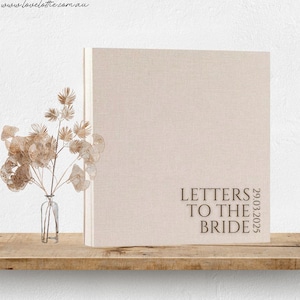 Pode incluir: Um livro bege coberto de linho com o título "Letters to the Bride" e a data 29.03.2025 impressa na capa. O livro é exibido em uma prateleira de madeira ao lado de um pequeno vaso de vidro com flores secas. O fundo é uma parede branca.