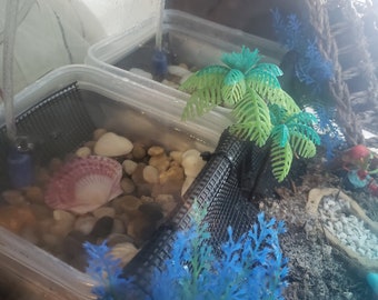 May include: A small aquarium setup with a clear plastic container. The tank features a black mesh filter, small pebbles, a pink seashell, and artificial blue and green plants. A small palm tree decoration is also present.