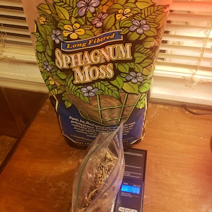 May include: A bag of long-fibered Sphagnum Moss, with floral illustrations, sits next to a clear plastic bag of moss on a digital scale. The scale displays a weight of 102.0 grams. The bag is on a wooden surface.