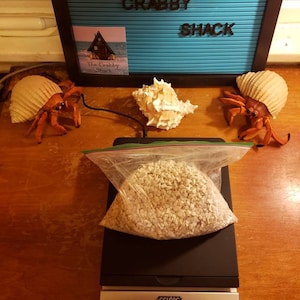 May include: A clear plastic bag filled with small, light-colored gravel sits on a black digital scale, displaying a weight of 1.806. The scale is labeled "Accuteck." Decorative hermit crabs and seashells surround a sign that reads "THE CRABBY SHACK."
