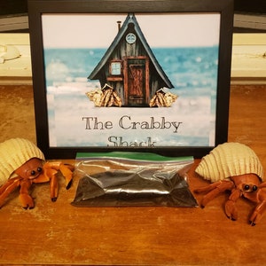 May include: A framed print reading 'The Crabby Shack' with two hermit crab ornaments and a bag of dark substrate. The print shows a cabin on a beach. The crabs have orange bodies and are in beige shells.