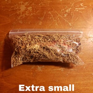 May include: A clear plastic bag filled with dried, brown plant material, labeled "Extra small" in white text. The bag is sealed and rests on a wooden surface with visible scratches.