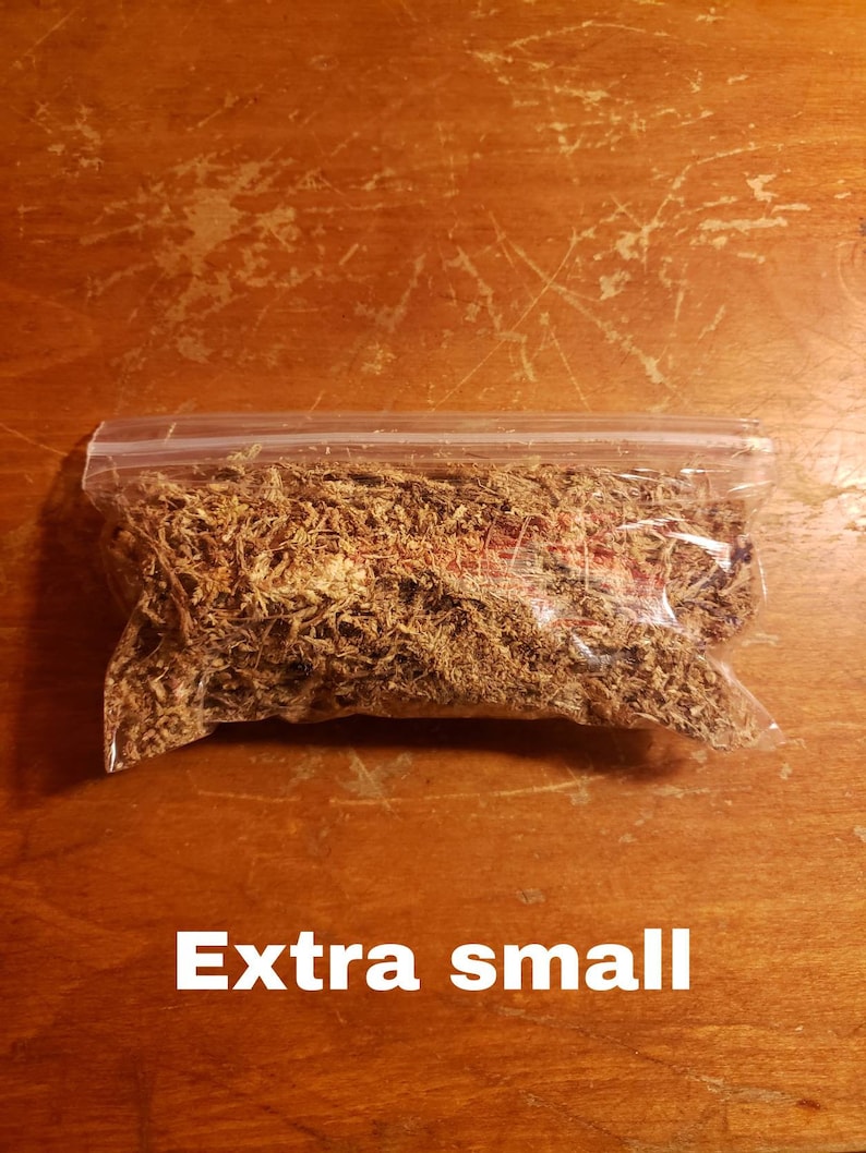 May include: A clear plastic bag filled with dried, brown plant material, labeled "Extra small" in white text. The bag is sealed and rests on a wooden surface with visible scratches.