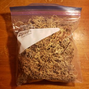 May include: A clear Ziploc bag filled with dried, brown plant matter, labeled "Medium" in white text. The bag is sealed with a blue zipper and sits on a wooden surface.