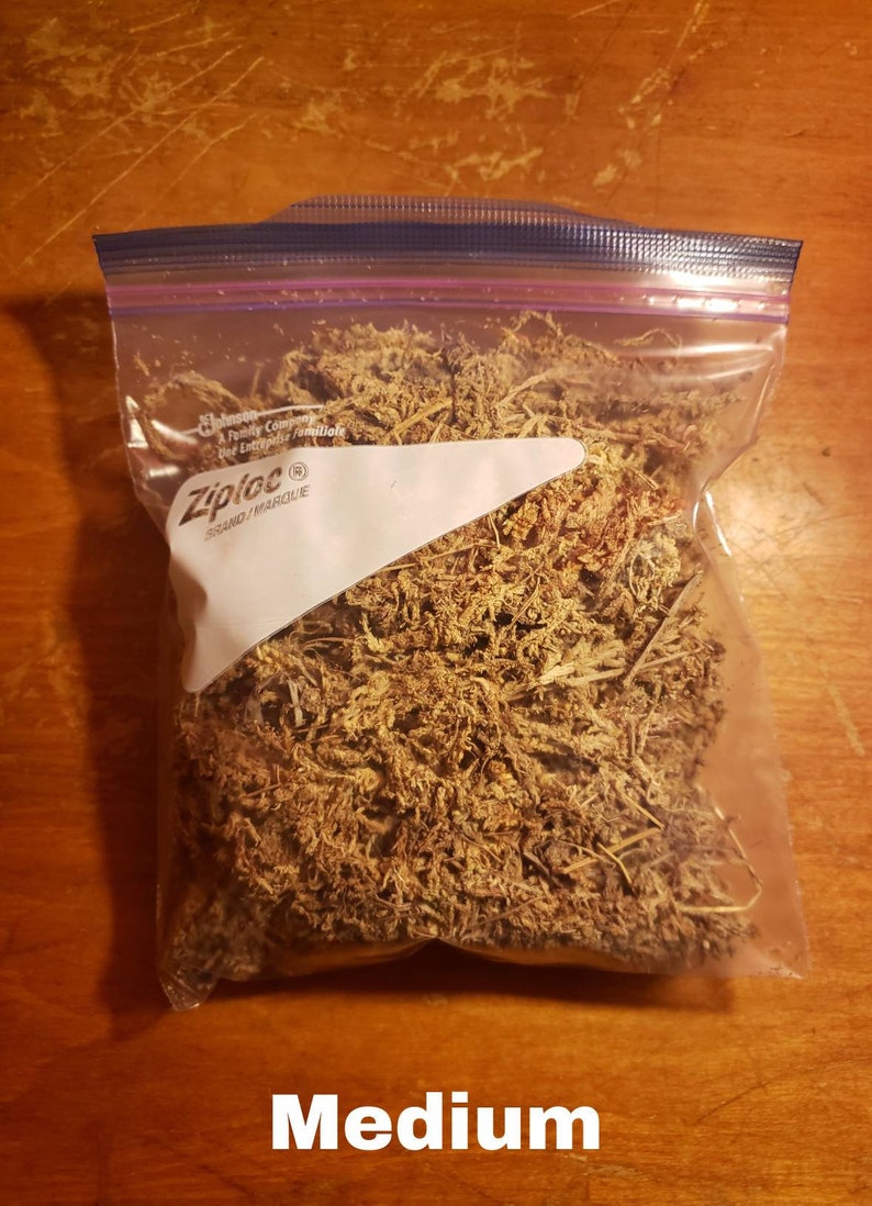 May include: A clear Ziploc bag filled with dried, brown plant matter, labeled "Medium" in white text. The bag is sealed with a blue zipper and sits on a wooden surface.