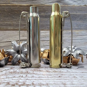 May include: Two bullet casing bottle stoppers, one silver and one gold, with wire clip handles. Decorative metal flower accents are scattered around the stoppers. The items are displayed on a weathered wooden surface.