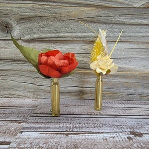 May include: Two floral boutonnieres. One features a red flower with a green leaf, the other has yellow and cream-colored flowers. Both are mounted on brass bullet casings, set against a rustic wood background.