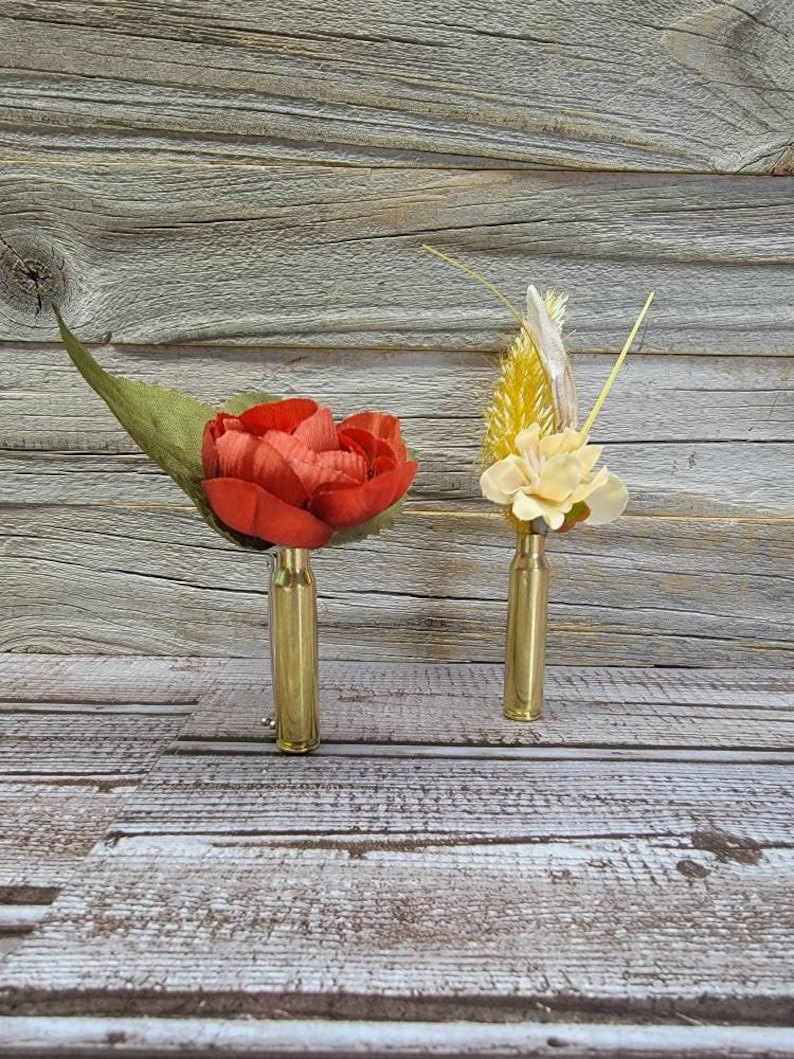 May include: Two floral boutonnieres. One features a red flower with a green leaf, the other has yellow and cream-colored flowers. Both are mounted on brass bullet casings, set against a rustic wood background.