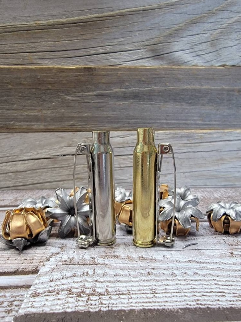 May include: Two bullet casing bottle stoppers, one silver and one gold, with safety pin handles. Several decorative bullet casings in silver and gold are scattered around the stoppers, all on a wooden surface.