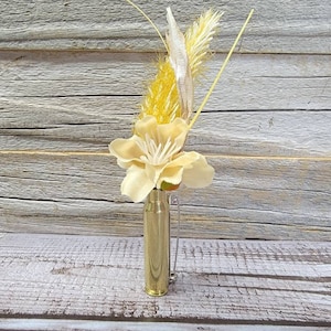 May include: A small, gold-colored bullet casing repurposed as a vase, holding a delicate arrangement of dried flowers and grasses. The arrangement includes a large, pale yellow flower and feathery, light yellow and beige accents. The vase is on a weathered wooden surface.
