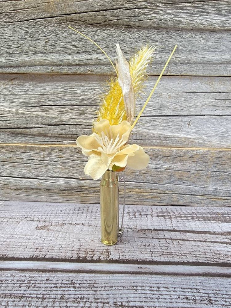 May include: A small, gold-colored bullet casing repurposed as a vase, holding a delicate arrangement of dried flowers and grasses. The arrangement includes a large, pale yellow flower and feathery, light yellow and beige accents. The vase is on a weathered wooden surface.