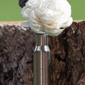 May include: A unique floral arrangement featuring a white flower and dried lavender sprigs in a repurposed bullet casing. The metallic casing contrasts with the natural elements, creating a rustic and artistic display. The arrangement is set against a blurred green background.