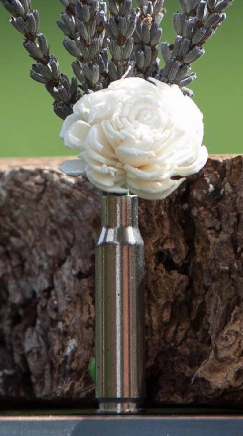 May include: A unique floral arrangement featuring a white flower and dried lavender sprigs in a repurposed bullet casing. The metallic casing contrasts with the natural elements, creating a rustic and artistic display. The arrangement is set against a blurred green background.
