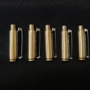 May include: Five brass bullet casings with silver safety pins attached, arranged on a black surface. The casings are cylindrical with a rounded base and a narrower top. The safety pins are straight with a simple clasp.