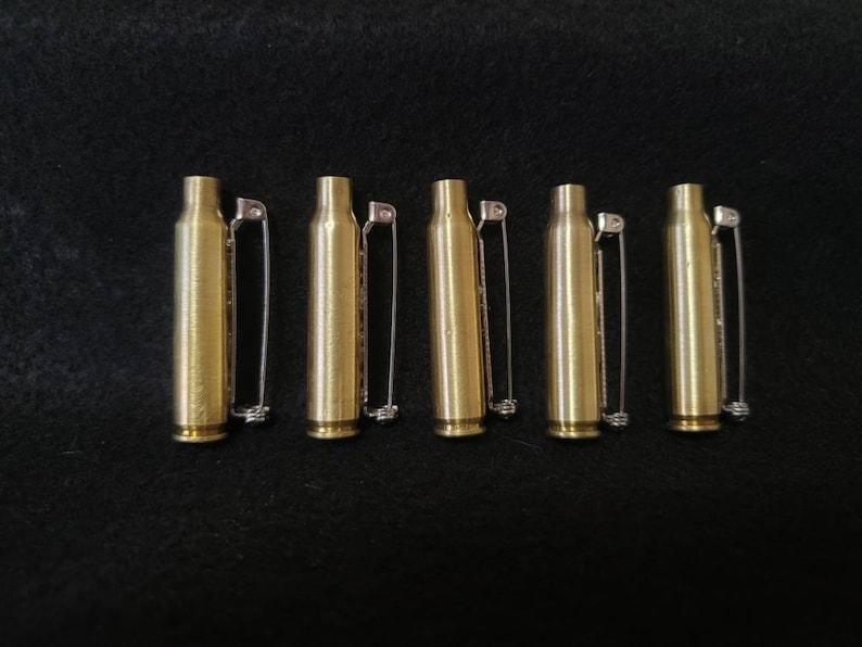 May include: Five brass bullet casings with silver safety pins attached, arranged on a black surface. The casings are cylindrical with a rounded base and a narrower top. The safety pins are straight with a simple clasp.