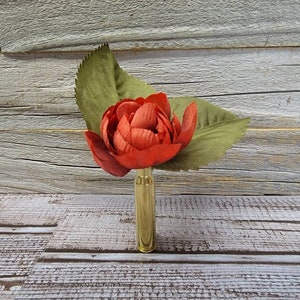 May include: A red fabric flower with green leaves is attached to a gold bullet casing. The flower is in full bloom, and the leaves are a muted green. The background is a weathered wood surface.