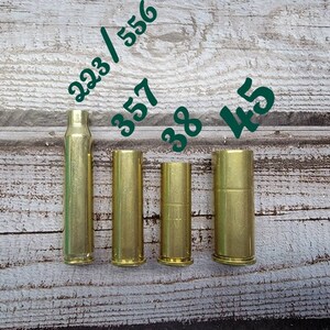 May include: Four brass bullet casings of varying sizes are displayed against a wood-grain background. The casings are labeled with the numbers 223/556, 357, 38, and 45 in green. The casings are empty and cylindrical, showcasing their different lengths.