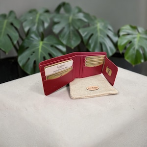 Può includere: Portafoglio in pelle rosso aperto con più fessure per carte, contenente carte con il logo "American Express". Un piccolo astuccio beige con un'etichetta in pelle è accanto al portafoglio.
