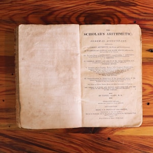 Puede incluir: Un libro antiguo abierto, titulado "The Scholar's Arithmetic" con "Federal Accountant" impreso debajo. Las páginas del libro están envejecidas y amarillentas, sobre una superficie de madera cálida con vetas y nudos visibles.