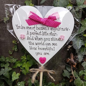 Pode incluir: Um letreiro de madeira branco em forma de coração com uma fita rosa e flores rosas. O letreiro diz "You're one of heaven's angels now, A perfect little star, And when you shine the world can see How beautiful you are."
