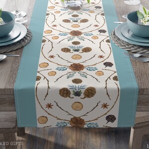 May include: A white table runner with a blue border features a repeating pattern of pine cones, branches, and star anise. The runner measures 16 inches by 72 inches.