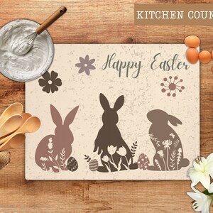 May include: A rectangular kitchen counter mat with a beige background and a distressed texture. The mat features three bunny silhouettes with floral designs, Easter eggs, and the words "Happy Easter". Decorative flowers and eggs are also present.