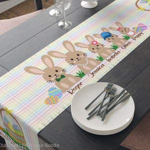 May include: A pastel pink, yellow, and blue checkered table runner with a row of brown bunnies with Easter eggs. The bunnies are decorated with bows, pearls, and a green bow tie. The names Roger, Jessica, Isabella, Dillon, and Poppy are written on the runner.