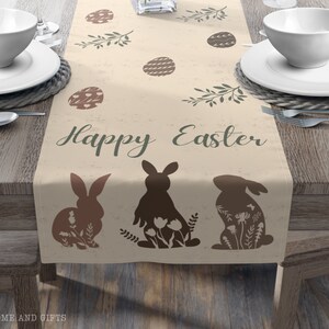 May include: A beige table runner with a brown and green Easter theme. It features the text "Happy Easter" and silhouettes of bunnies with floral designs.