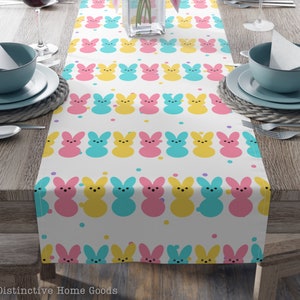 May include: A white table runner with a repeating pattern of pastel pink, yellow, and blue bunny shapes. The bunny shapes are outlined in black and have a simple, cartoon-like design. The background is white with small, scattered dots of various colors.