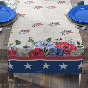 May include: A white linen table runner with a red, white, and blue border featuring stars. The runner is decorated with a floral pattern of red, white, and blue flowers.
