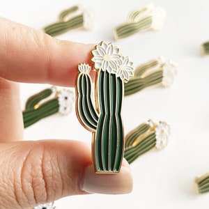 May include: A gold-tone enamel pin featuring a green cactus with white flowers. The cactus has a detailed design with multiple stems and a rounded top.