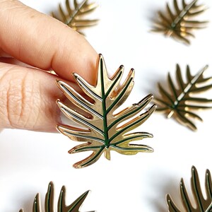 May include: Gold-colored metal pin with a green leaf design. The leaf has a detailed vein pattern.