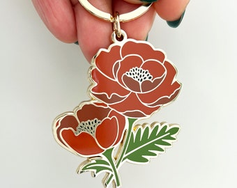 May include: Gold-tone keychain featuring two red poppy flowers with green leaves. The flowers have white centers with black accents.