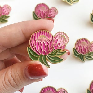 May include: A vibrant enamel pin featuring a protea flower design. The pin showcases a bright pink flower with green leaves and gold trim. The flower is held in a hand, with other pins visible in the background.
