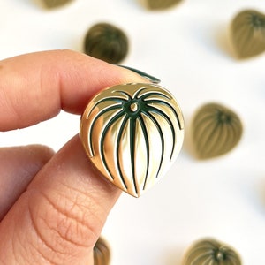 May include: Gold-colored heart-shaped pin with a green leaf design. The pin has a raised, textured surface.