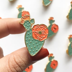 May include: A gold-tone enamel pin featuring a green cactus with orange flowers. The cactus has a heart shape and is decorated with small white dots.
