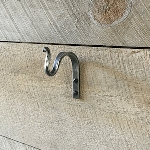 Może przedstawiać: Srebrny metalowy hak z ozdobnym zwojem na górze, zamontowany na jasnej drewnianej ścianie.
